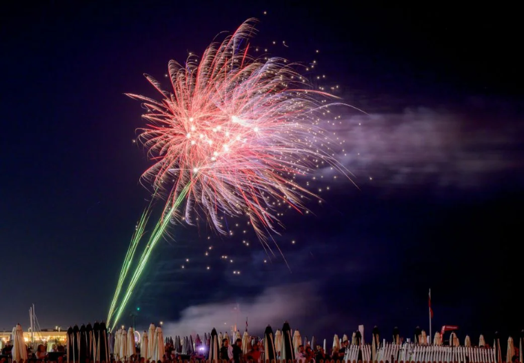 Colorful fireworks explode over a beach at night as a crowd watches from behind rows of umbrellas and tents.