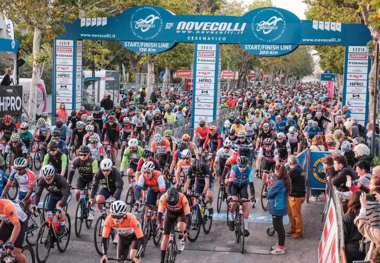 Massive peloton of cyclists at the start line beneath a blue arch with ‘Novecolli’ logos, spectators lining both sides.