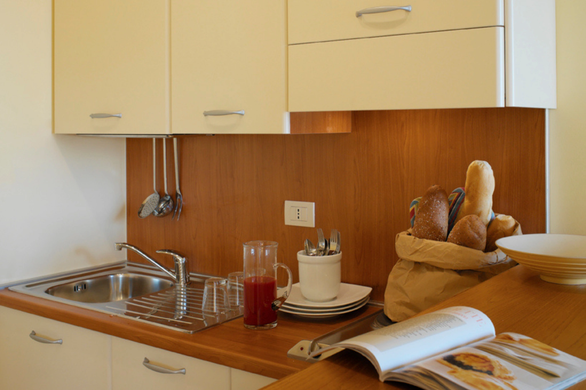 La kitchenette di un miniappartamento: lavello, piano di lavoro in legno, pane, stoviglie e un ricettario aperto